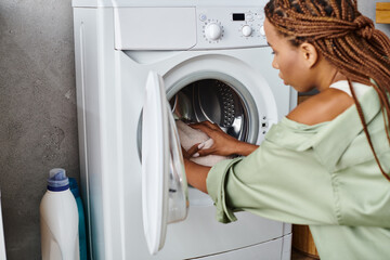 African American woman with afro braids putting clothes into a dryer in a bathroom while doing...