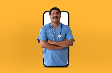 A cheerful Indian man doctor wearing blue scrubs and a stethoscope stands with arms crossed,...