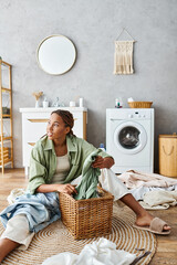 African American woman with afro braids sitting on the bathroom floor with a laundry basket, doing...