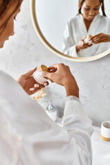 An African American woman with afro braids in a bathrobe holding beauty jar with cream in front of...