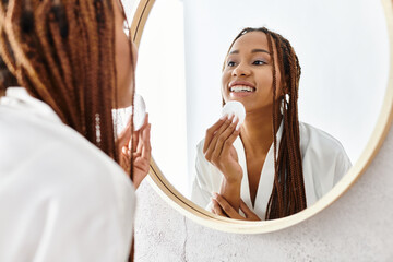 An African American woman with afro braids in a bathrobe using cotton pad in front of a mirror in a...