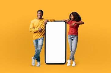 A cheerful black couple stands to the left and right of a large smartphone mockup with a blank...