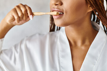 Afro braided African American woman in bathrobe brushes teeth with a wooden toothbrush in modern bathroom.