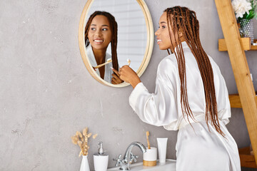 An African American woman with afro braids in a bathrobe brushing her teeth in a modern bathroom in...