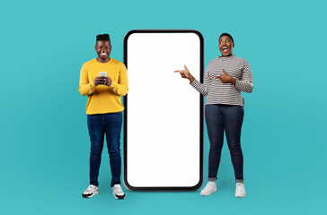 Two smiling black people interact with their mobile devices while standing on a turquoise...