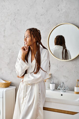 African American woman with afro braids stands in bathrobe before the modern bathroom mirror.