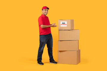 A cheerful delivery worker dressed in a red uniform points towards a stack of three cardboard boxes...