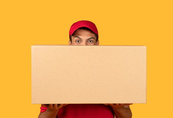 A delivery man wearing a red uniform and cap is holding a large cardboard box. The background is a...