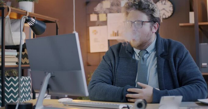 Loop of male office worker inhaling and exhaling smoke from e-cigarette sitting at computer desk at work and looking at monitor. Smoking and workplace concept.