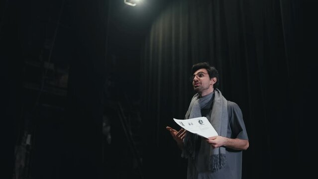 Side view of a confident male stage director in a gray T-shirt and scarf walking onto the stage along with a stack of papers and greeting everyone in the theater with black curtains