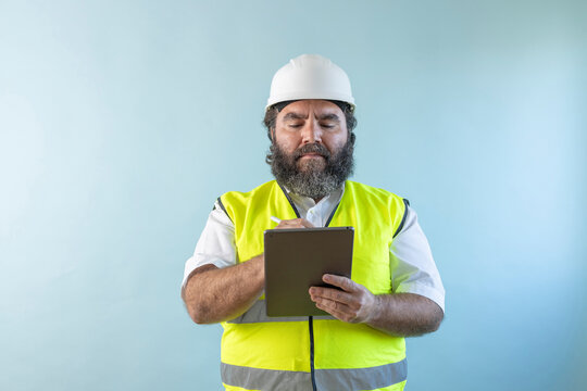 serious adult man engineer with beard and wearing helmet and safety vest looking the touch pad on blue background, using his tablet and reviewing work