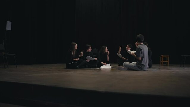 A group of young confident actors in black uniforms are talking with the director about the script for their next play and are sitting on stage in the theater