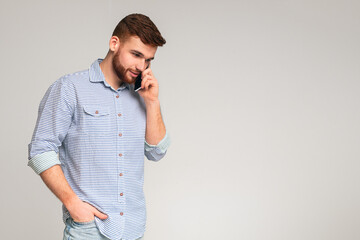Handsome young bearded man talking on cellphone and looking away on grey studio background, copy...