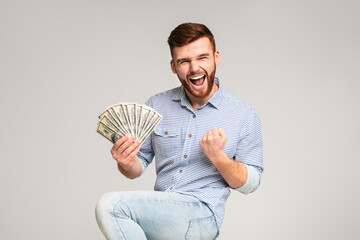 Win a lottery. Young handsome guy happy to have fan of dollars in hand, grey studio background,...