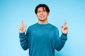 Asian young guy stands against a blue background, with his eyes closed and fingers crossed,...