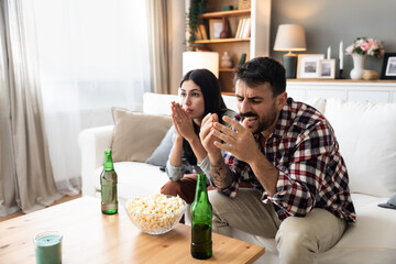 Excited and tense young couple sitting on sofa at home watching or streaming sports event on tv...