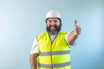 smiling adult man engineer with beard and wearing helmet and safety vest looking at camera on blue background, making thumb up