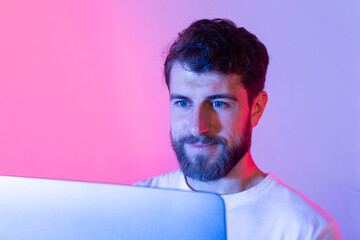 A man with a beard sits in front of a laptop computer, typing and browsing the internet. He is...