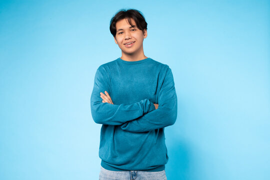 Asian guy stands with his arms crossed in front of a solid blue background. He appears confident and assertive in his posture. The blue background creates a stark contrast