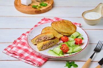Potato pancakes stuffed with minced meat on a ceramic plate on a light wooden background. Served with fresh cucumber and cherry tomatoes. Belarusian cuisine.
