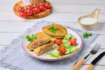 Potato pancakes stuffed with minced meat on a ceramic plate on a light wooden background. Served with fresh cucumber and cherry tomatoes. Belarusian cuisine.