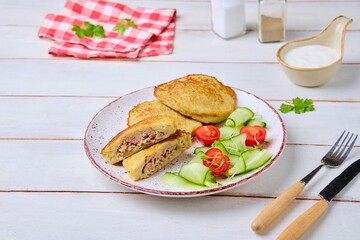 Potato pancakes stuffed with minced meat on a ceramic plate on a light wooden background. Served with fresh cucumber and cherry tomatoes. Belarusian cuisine.