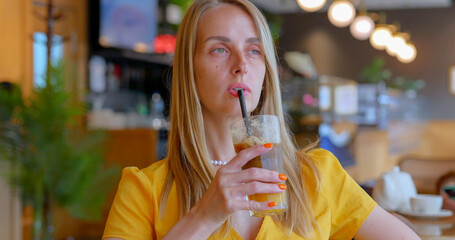 A blonde girl drinks iced coffee with orange juice in a cozy cafe