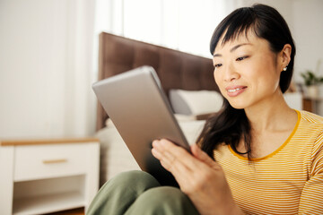 A happy asian woman sitting on a bedroom floor and smiling at the tablet.
