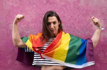 Trans woman in strong arm pose with rainbow flag isolated on pink background and looking at camera