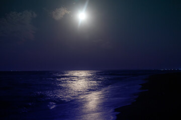 Sea in the moonlight. The moon is shining above the sea water, lights of the village on the horizon