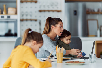 Fototapeta premium A hard-working mother having a video call on her laptop while working from home and looking after her kids