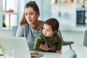 A chagrined mother holding her baby as the child curiously touches the laptop and disrupts mother's work making her feel stressed