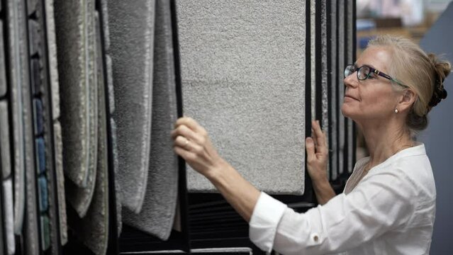 Smiling mature woman touching carpet looking through samples for home redecorating project, in a floor store.