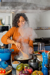 Hispanic young woman standing holding wooden spoon mixing food in frying pan while cooking in kitchen at home