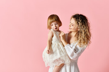 Mother in white dress holding girl in white dress on pink background.