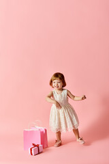 Little girl in white dress with pink gift bags against pink backdrop.