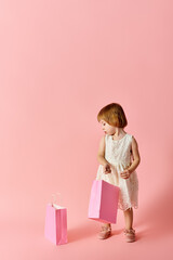 Adorable girl in white dress holding shopping bags on pink background.
