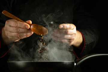 Adding dry pepper to a hot frying pan with a spoon in the cook hand. Cooking concept on black...