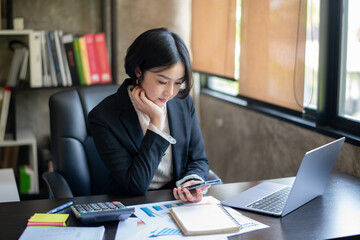 A woman in a business suit is sitting at a desk with a laptop and a cell phone