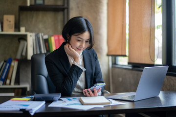 A woman in a business suit is sitting at a desk with a laptop and a cell phone