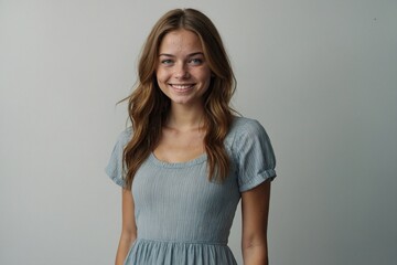 Obraz premium A portrait of a young woman with natural makeup, wearing a casual grey dress, looking directly at the camera with a happy expression against a plain grey background.