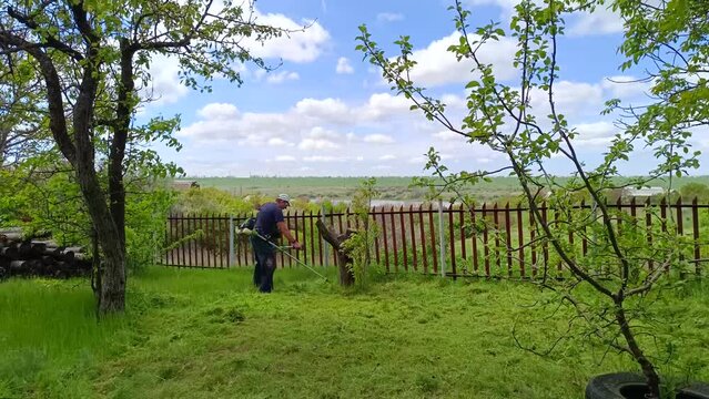 A man in a cap with a petrol scythe-trimmer mows bright green grass near the fense
 on a garden plot on a sunny spring day against the backdrop of the blue sky, sea estuary, woodpil, fields