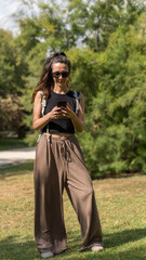 A woman is sitting on grass, using a laptop and drinking coffee with modern buildings in the background