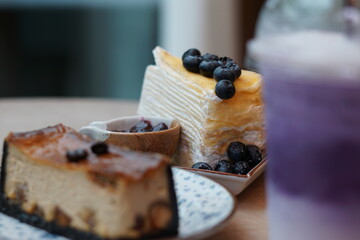 A piece of blueberry crepe cake topped with blueberry on the plate with blur of espresso macadamia cheese cake and sweet potato milk tea.