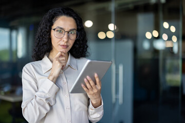 A professional Hispanic woman with curly hair and glasses is focused on her task, using a tablet in...