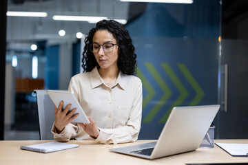 A professional woman in an office environment wearing glasses, focused and utilizing a tablet...