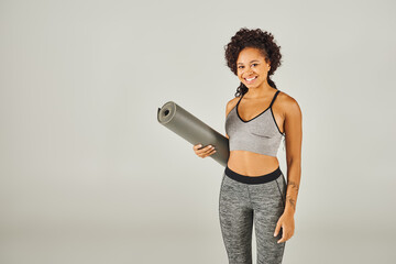A curly African American sportswoman in activewear holding a yoga mat in a studio with a grey...