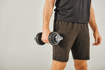 A young sportsman in active wear energetically lifts a pair of dumbbells in a studio with a grey...