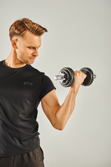 A young sportsman in active wear showcases his strength, holding a pair of dumbbells in a studio with a grey background.