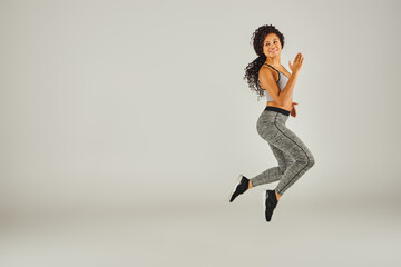 A young African American woman in athletic attire joyfully jumps against a plain gray backdrop in a studio setting.